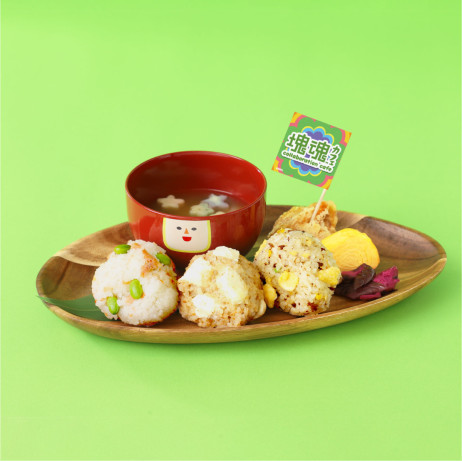 A wooden plate featuring three round onigiri with a red bowl of miso soup with star shapes inside. The bowl of miso soup has a face on it (it's Prince's cousin Miso).