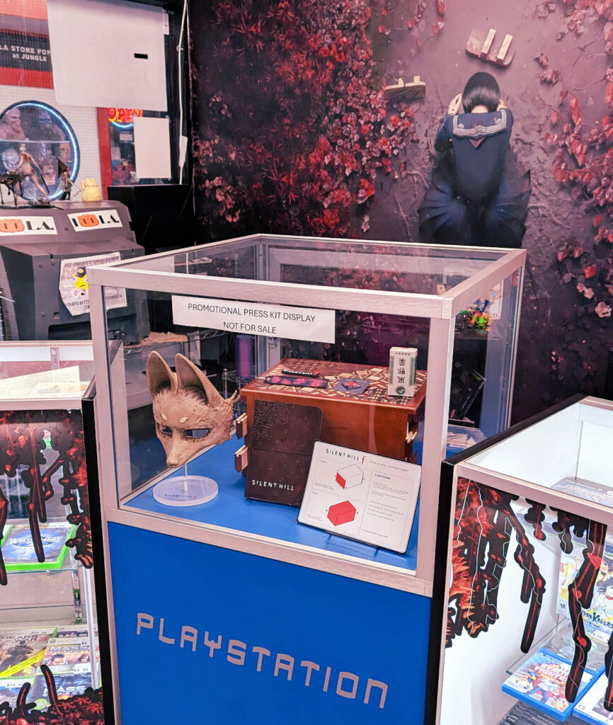 Photo of Silent Hill f PR box including a fox mask and puzzle box in a glass case and a decal of the Silent Hill protagonist on a flowery background behind